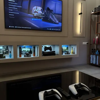 Wartebereich mit Fernseher, beleuchteten Regalen und zwei PS5-Controllern